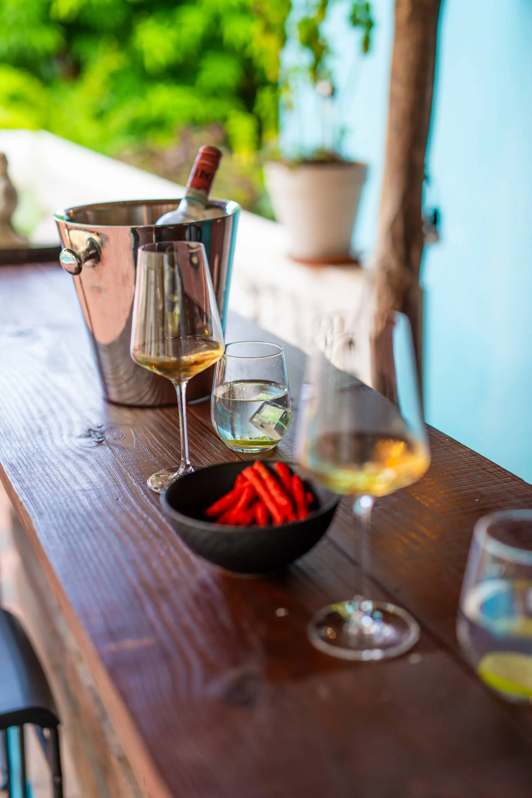 A stylish outdoor bar setup at Casa Leonore in Curaçao, featuring a chilled bottle of wine in an ice bucket, two wine glasses, a glass of water with lime, and a bowl of vibrant red chili peppers, creating a cozy and inviting atmosphere.